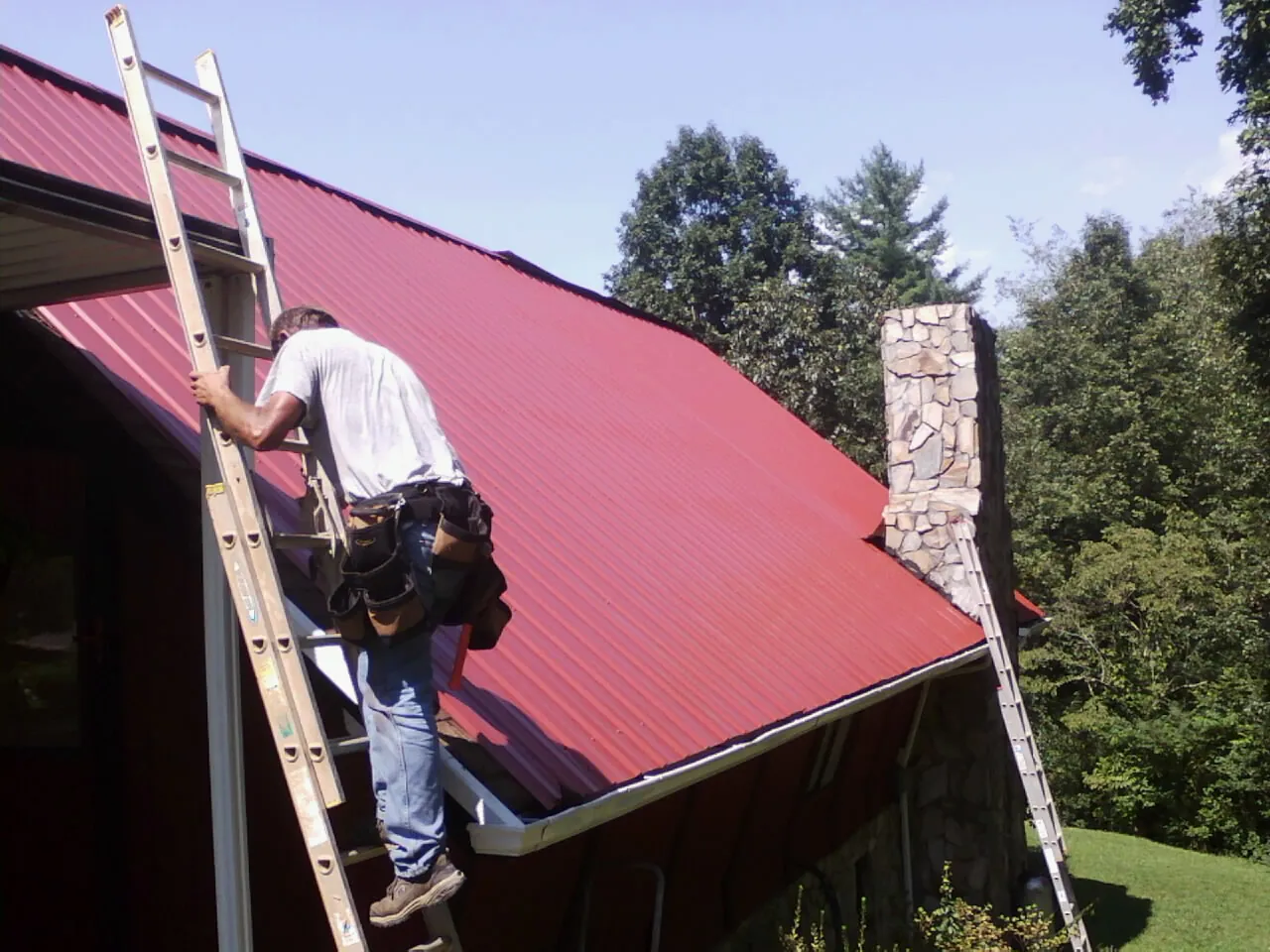 Roofer on a ladder beside a red metal roof and stone chimney — Soffit & Fascia Repair by Roofing Brothers in Ketchum